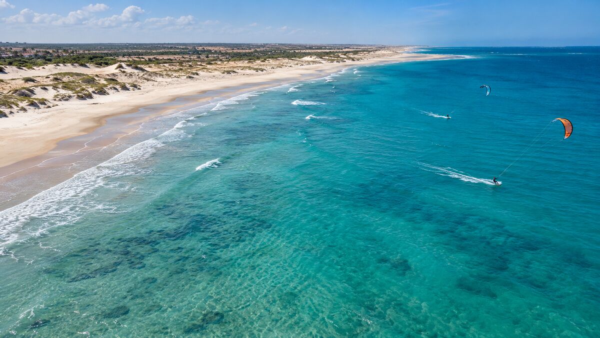 Vista aérea de praia com dunas, mar azul-turquesa e kitesurfistas